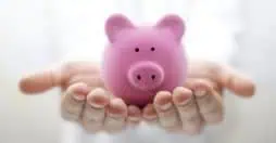 Dos manos sostienen con suavidad una alcancía rosa contra un fondo suave y difuso. La imagen transmite temas de ahorro, cuidado y seguridad financiera.