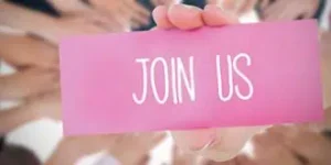 A hand holds a pink card reading "Join Us" in white letters, surrounded by a blurred circle of diverse hands reaching inward, evoking invitation and unity.