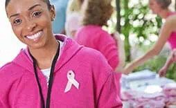 Mujer sonriente con un suéter rosa y un lazo de concientización sobre el cáncer de mama. El fondo muestra una mesa con personas vestidas de rosa, sugiriendo un evento de apoyo.