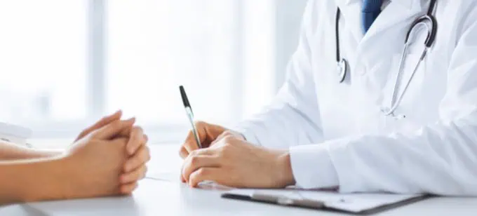 Un médico con bata blanca está sentado en un escritorio, con un bolígrafo y un portapapeles en la mano, hablando con un paciente cuyas manos entrelazadas descansan sobre la mesa. El ambiente es tranquilo y profesional.
